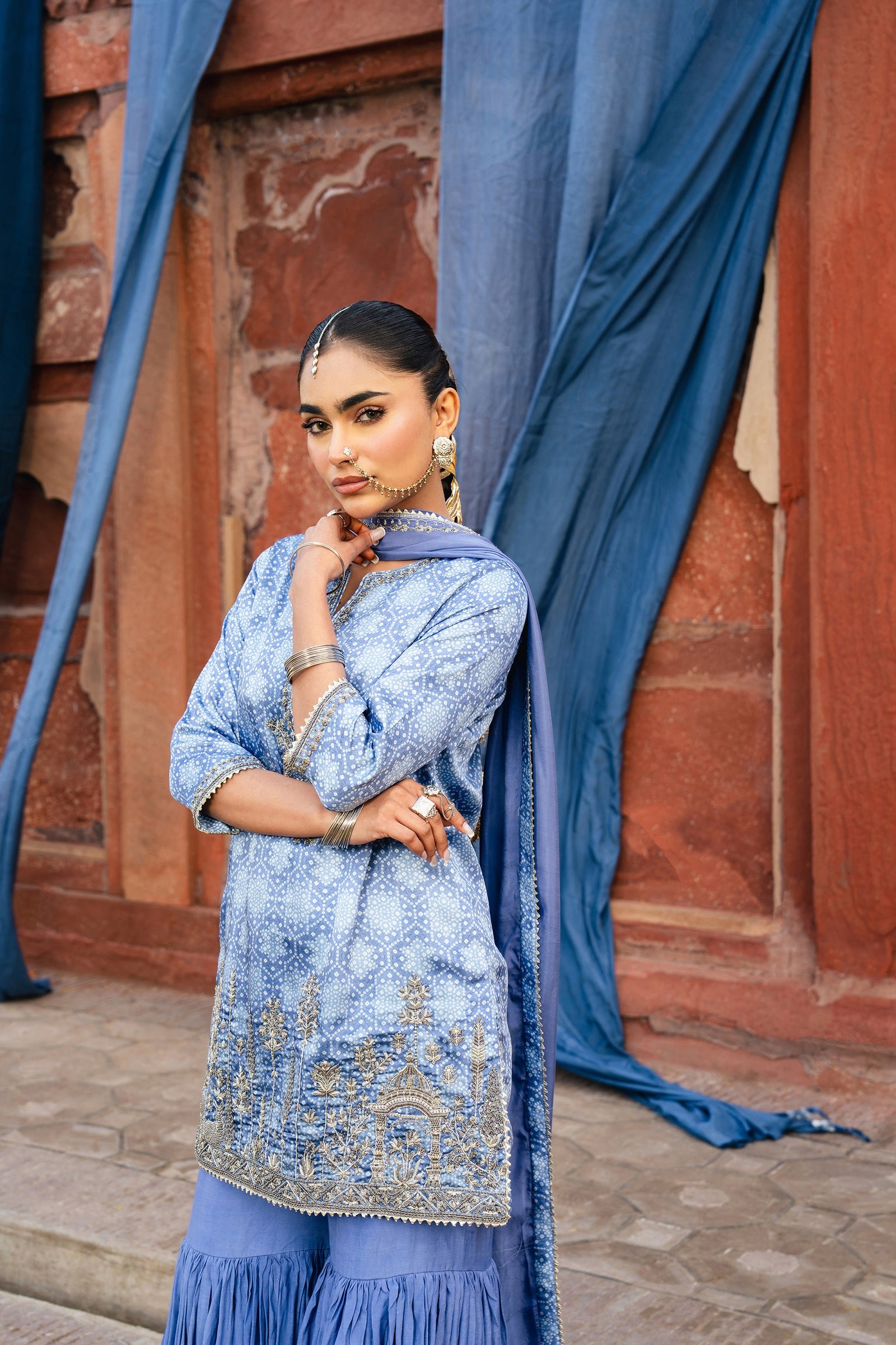 Model in a blue embroidered Anaa Khan kurta and sharara against a red sandstone wall with blue drapes